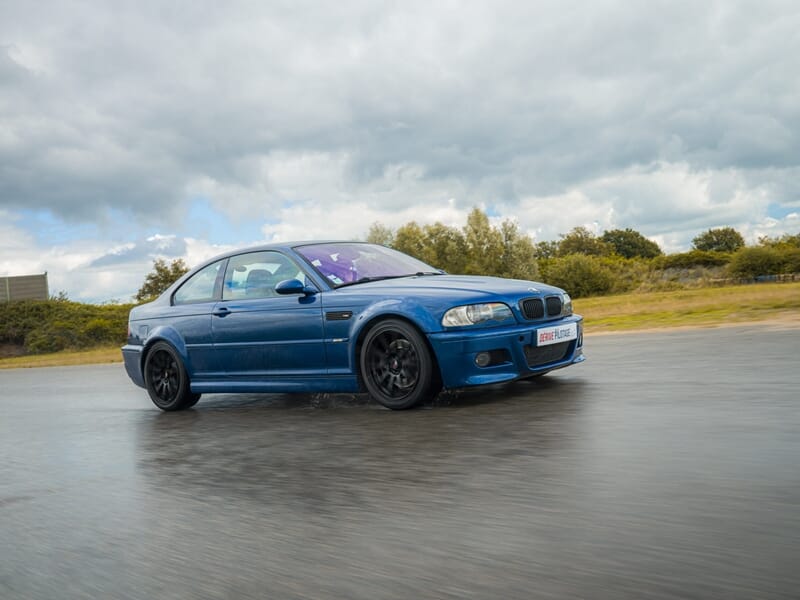 Voiture BMW bleue roulant sur circuit mouillé avec paysage vert et ciel nuageux lors d'un stage de pilotage glisse sur circuit.
