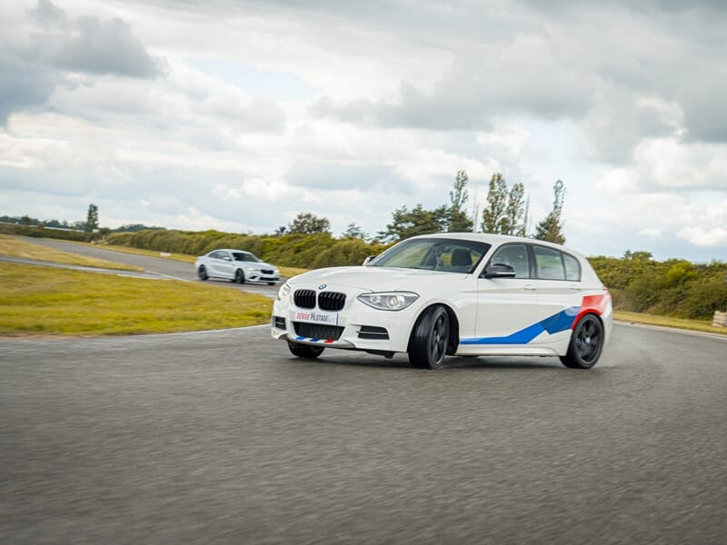 Voiture BMW blanche avec bandes colorées roulant sur circuit, avec une autre voiture en arrière-plan lors d'un stage de pilotage glisse sur circuit.