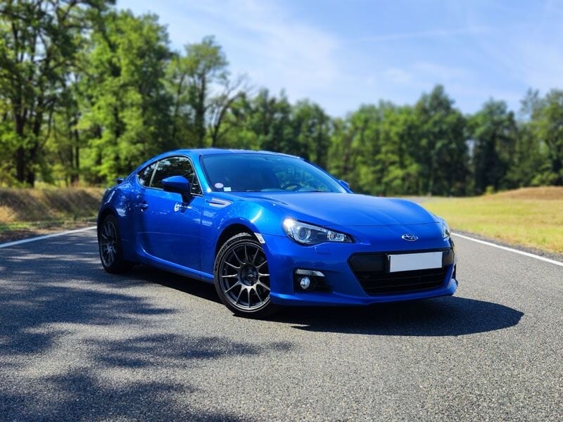 Voiture de sport bleue garée sur route entourée d'herbe et d'arbres, vue avant avec jantes noires.