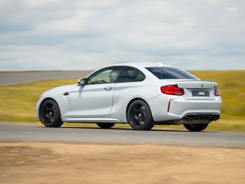 Voiture blanche sportive roulant sur un circuit de course avec un paysage de champs et ciel nuageux en arrière-plan.