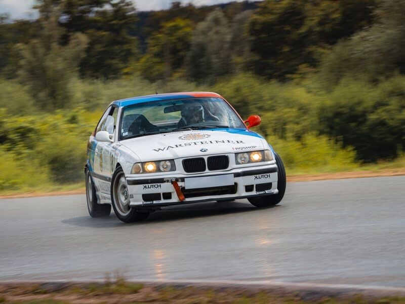 Voiture de course blanche en virage sur circuit, avec pilote concentré, entourée d'arbres verts en arrière-plan.