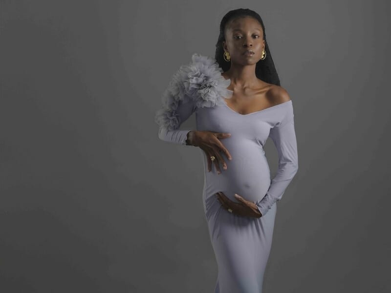 Femme enceinte en robe longue violette avec épaule dénudée, posant de face sur fond gris lors d'un shooting photo grossesse à Colmar.