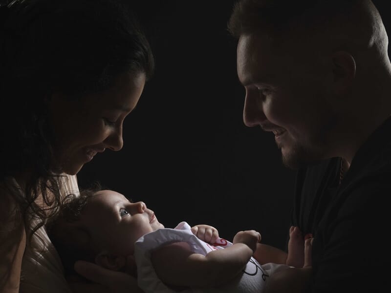 Un couple regarde avec tendresse un bébé dans leurs bras, fond noir lors d'un shooting photo famille à Colmar.
