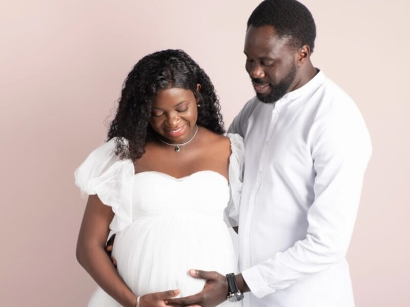Couple souriant en studio, la femme enceinte en robe blanche est entourée par son partenaire lors d'un shooting photo grossesse à Besançon.