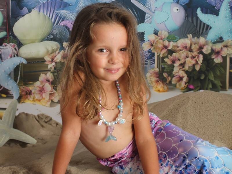 Petite fille déguisée en sirène avec une queue colorée et un collier, posant sur un décor marin avec coquillages et fleurs.