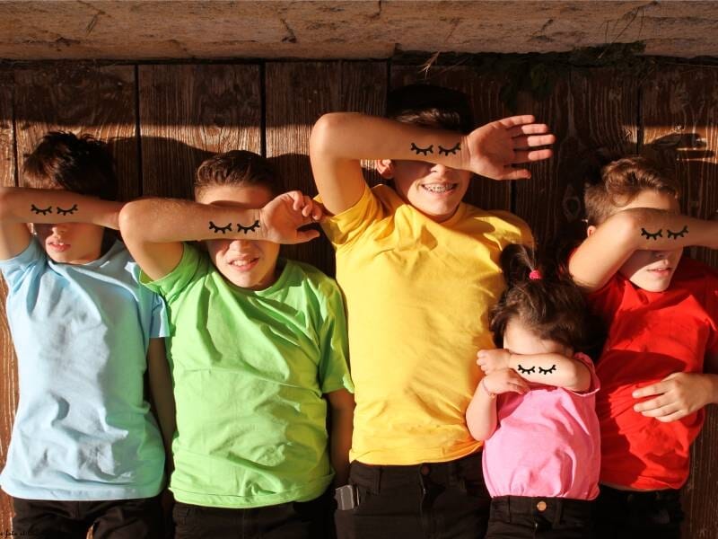 Cinq enfants avec des t-shirts colorés, allongés sur un sol en bois, bras levés avec dessins de cils sur les avant-bras, lors d'un shooting photo près d'Arles.
