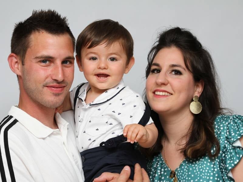 Portrait d'une famille souriante avec un père, une mère et leur jeune enfant dans un studio photo avec fond gris.