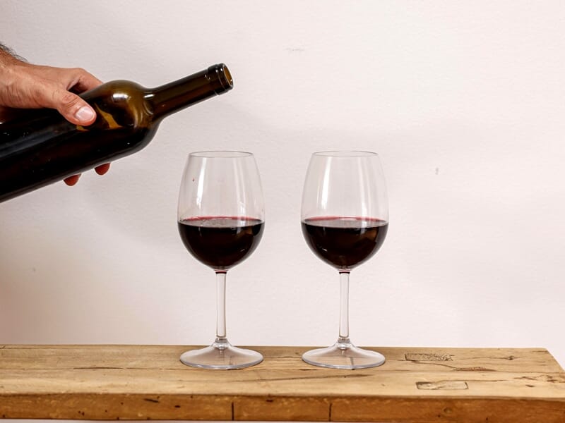 Main versant du vin rouge dans un verre à côté d'une bouteille et de raisins sur table.