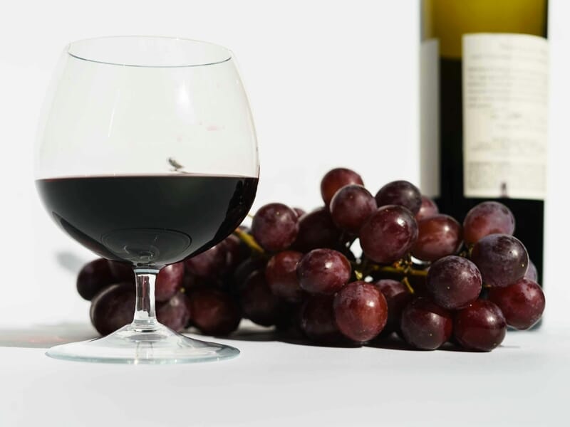 Verre de vin rouge à moitié rempli posé à côté d'une grappe de raisins rouges et d'une bouteille floue en arrière-plan.