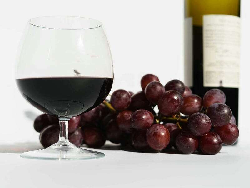 Verre de vin rouge à moitié rempli posé à côté d'une grappe de raisins rouges et d'une bouteille floue en arrière-plan.
