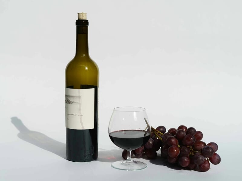 Bouteille de vin rouge avec bouchon, verre partiellement rempli et grappe de raisins rouges sur fond blanc