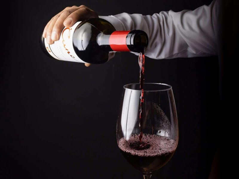 Un sommelier verse du vin rouge dans un verre à vin sur un fond noir, mettant en valeur la couleur et la texture du vin.