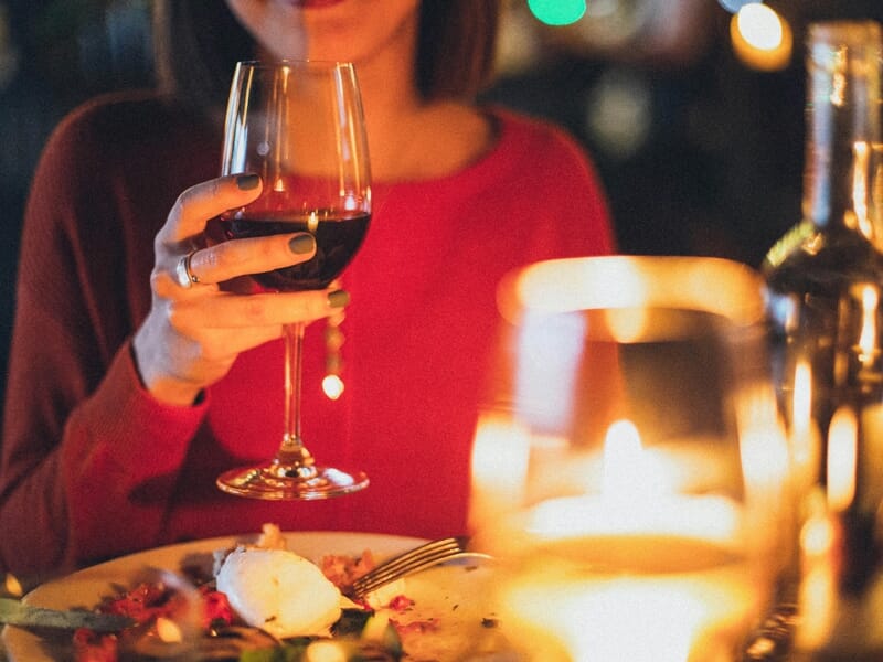 Une personne en pull rouge tenant un verre de vin rouge lors d'un dîner aux chandelles avec une assiette de nourriture devant elle.