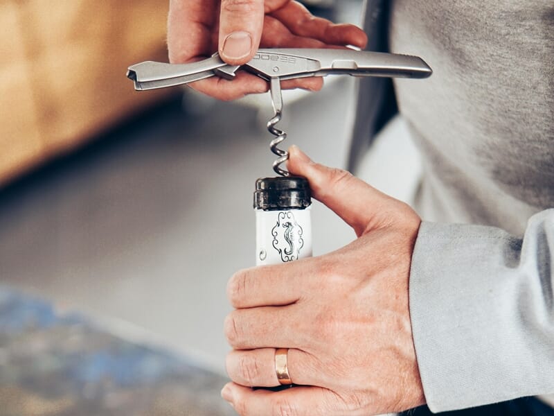 Une personne utilise un tire-bouchon en métal pour ouvrir une bouteille de vin avec une étiquette blanche décorée.