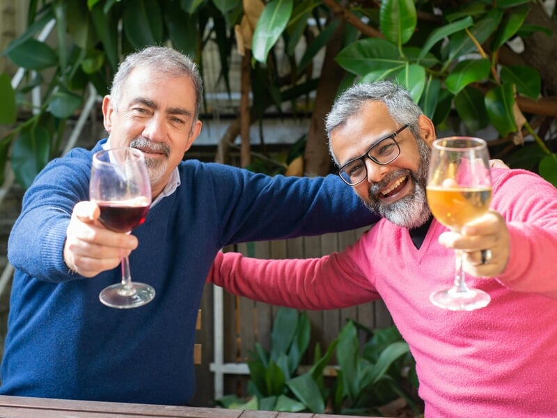 Deux hommes souriants trinquent avec un verre de vin rouge et un verre de vin blanc en extérieur, ambiance conviviale.