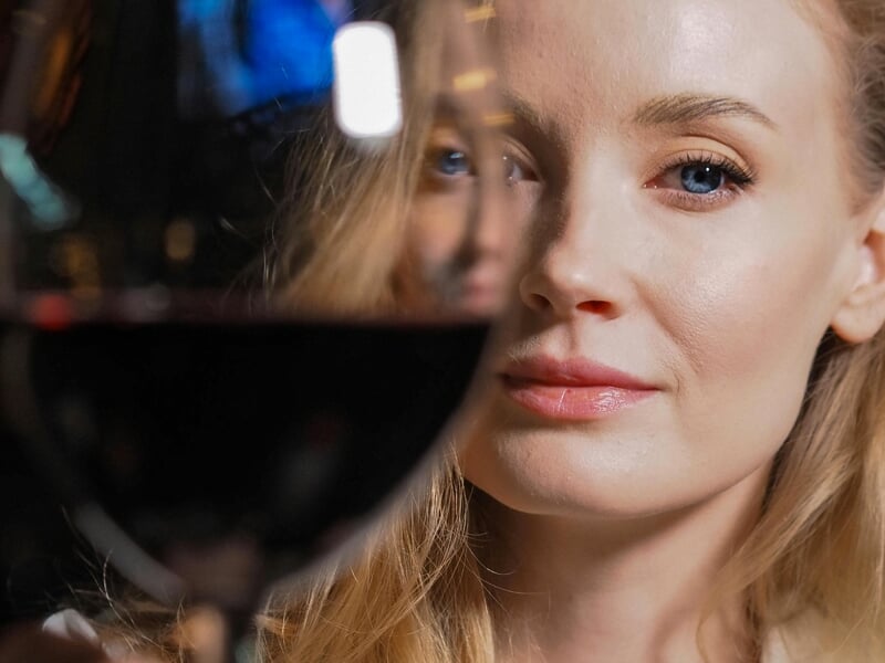 Femme blonde aux yeux bleus regardant à travers un verre de vin rouge, ambiance chaleureuse et élégante.