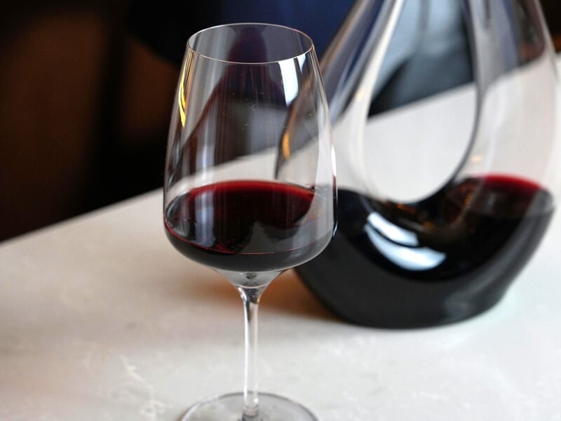 Verre de vin rouge à moitié rempli posé sur une table blanche avec une carafe en verre contenant du vin en arrière-plan.