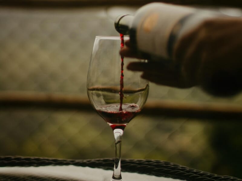 Un verre à vin est rempli de vin rouge versé d'une bouteille tenue par une main dans un cadre flou en arrière-plan.