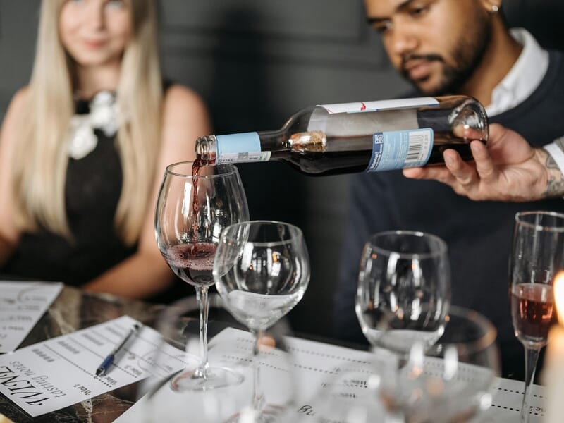 Un homme verse du vin rouge dans un verre lors d'une dégustation de vin, une femme blonde est assise en arrière-plan.