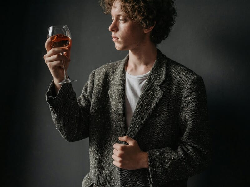 Jeune homme aux cheveux bouclés tenant un verre de vin rosé, observant la couleur et la texture avec attention.