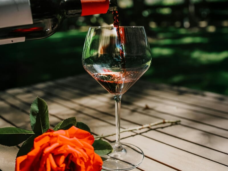 Un verre à vin est rempli de vin rouge versé d'une bouteille, posé sur une table en bois avec une rose orange à côté.