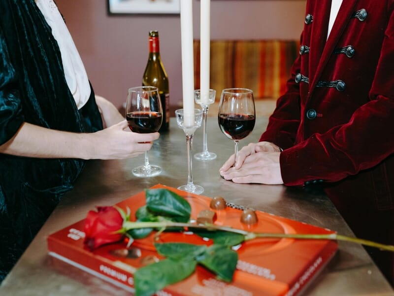 Deux personnes discutent autour d'un verre de vin rouge avec une rose rouge et une boîte de chocolats sur la table en métal.