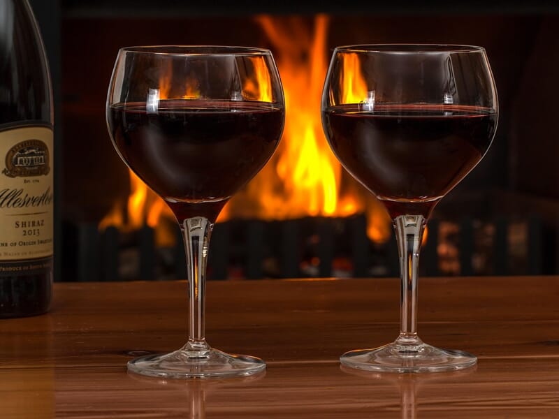 Deux verres de vin rouge posés sur une table en bois devant une cheminée allumée avec une bouteille de vin à gauche.