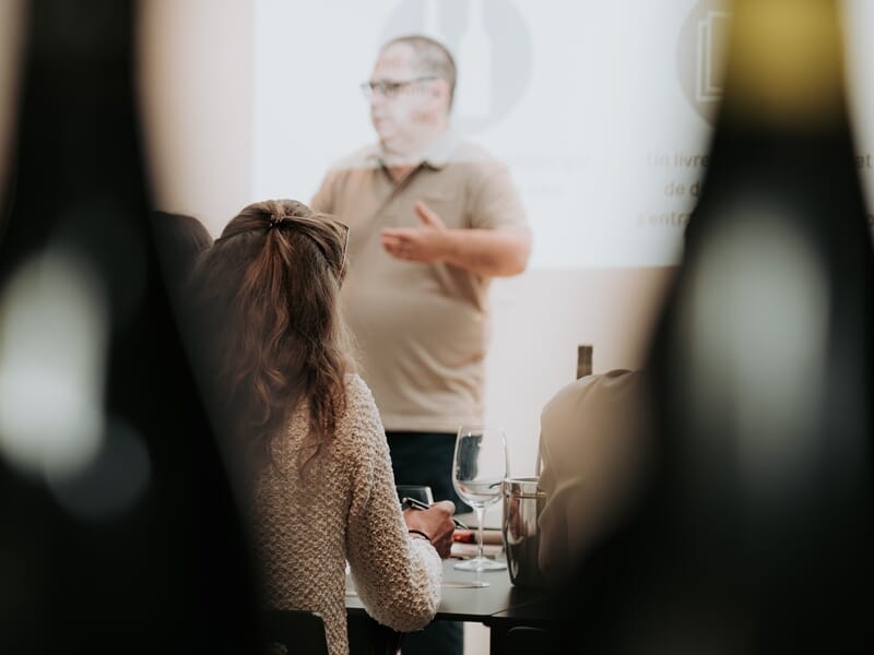 Personne assise de dos écoutant un homme qui donne une présentation sur l'œnologie avec un verre de vin à table.