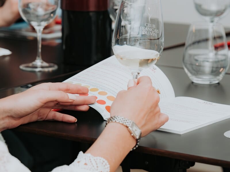 Personne tenant un verre de vin blanc lors d'une dégustation avec un guide des arômes sur une table en bois foncé.