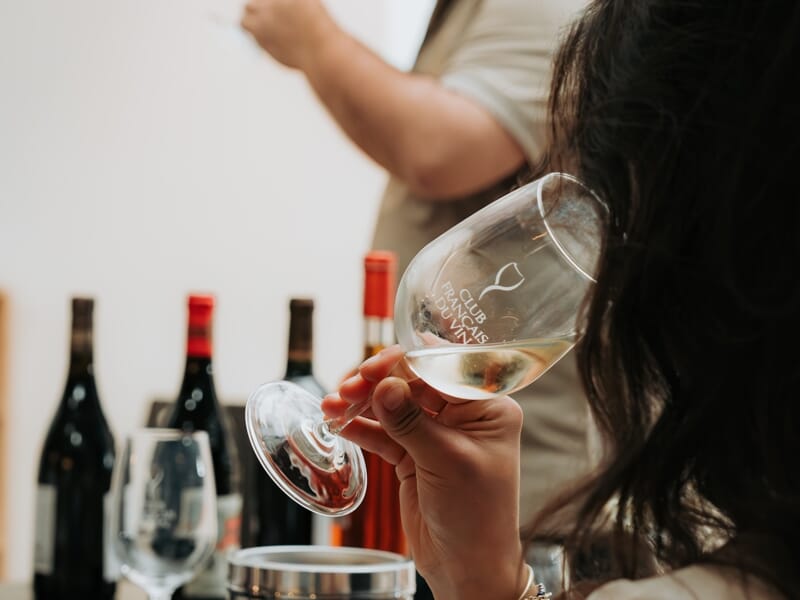 Personne dégustant un verre de vin blanc lors d'une séance d'oenologie avec plusieurs bouteilles en arrière-plan.