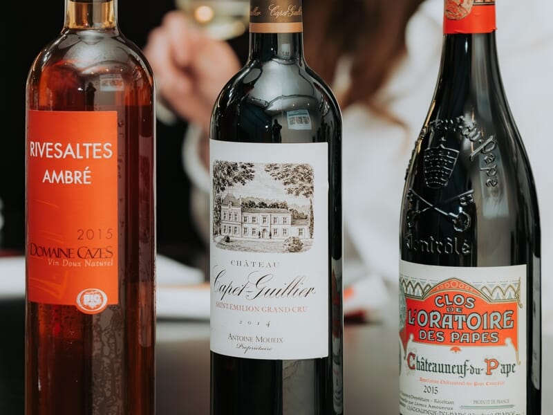 Trois bouteilles de vin différentes posées sur une table, avec des étiquettes visibles et une personne floue en arrière-plan.