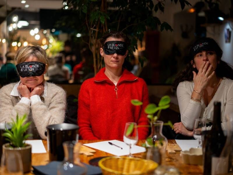 Trois personnes assises à une table, portant des bandeaux sur les yeux, dans une ambiance chaleureuse et conviviale.