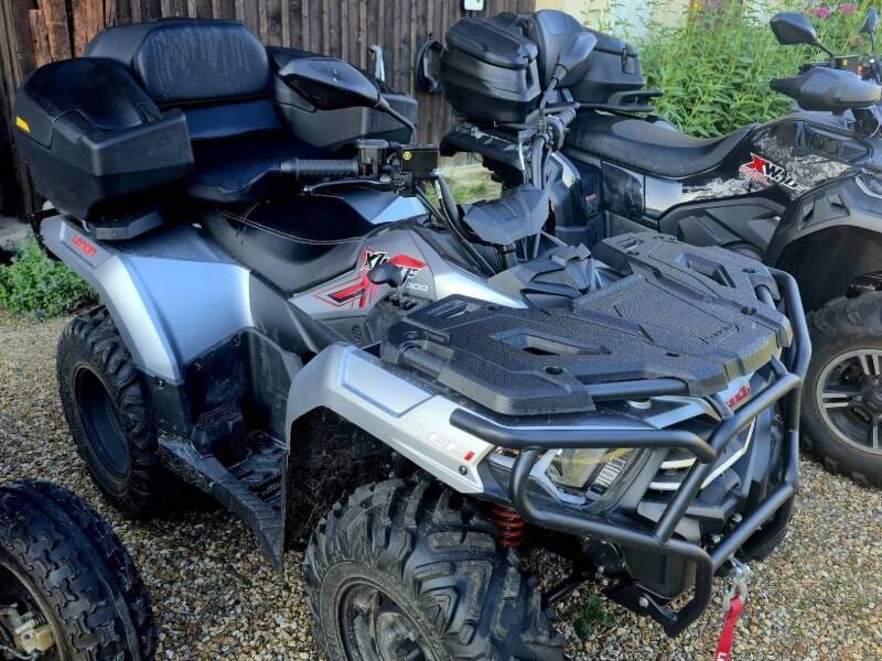 Quad tout-terrain gris avec siège avant et arrière noirs garé sur un sol en gravier.