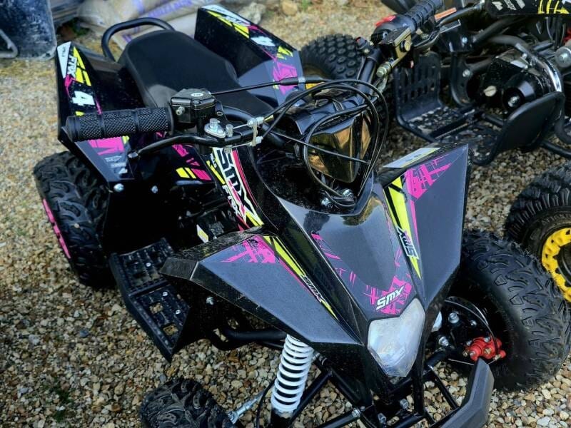 Quad noir avec des motifs roses et jaunes, posé sur un sol en gravier, adapté aux enfants.