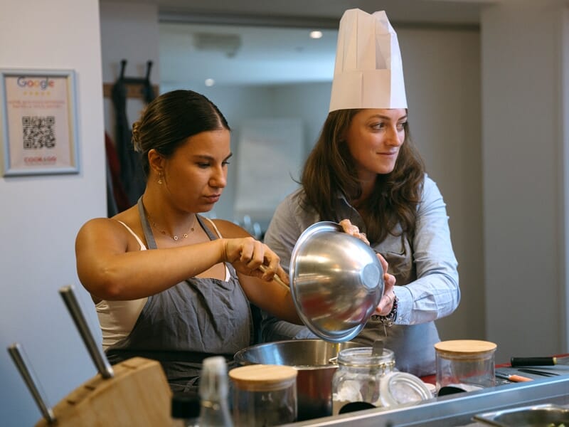Deux femmes en tablier participent à un cours de cuisine, l'une verse un ingrédient dans un saladier.
