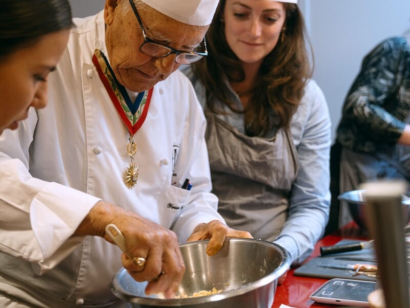 Un chef cuisinier âgé montre une technique de cuisine à deux femmes attentives dans un atelier culinaire.