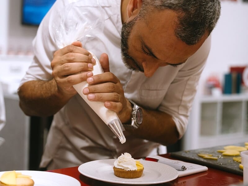 Un homme décore minutieusement une pâtisserie avec une poche à douille lors d'un cours de cuisine.