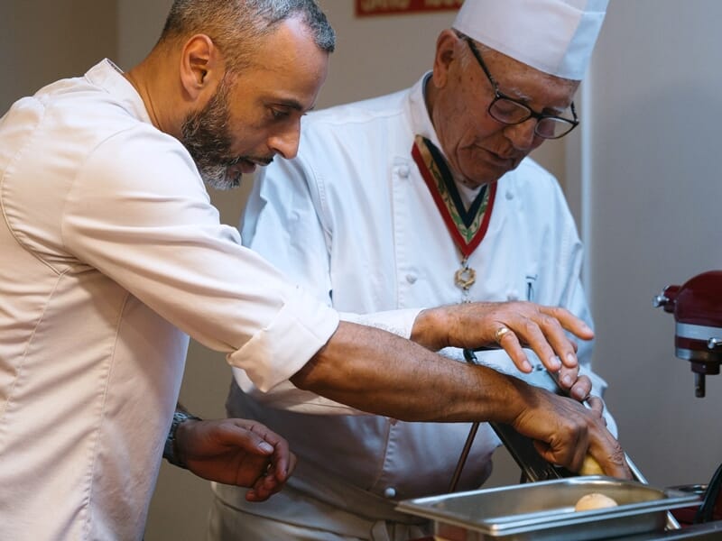Un chef expérimenté guide un homme dans la préparation d'un plat dans une cuisine professionnelle.