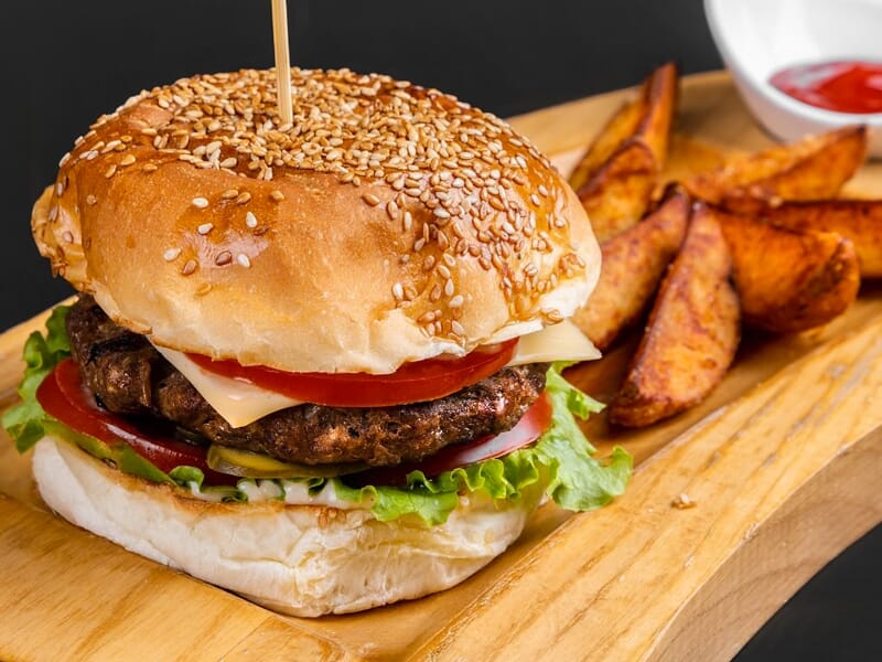 Un hamburger avec steak, fromage, tomate, salade et frites sur une planche en bois, prêt à être dégusté.