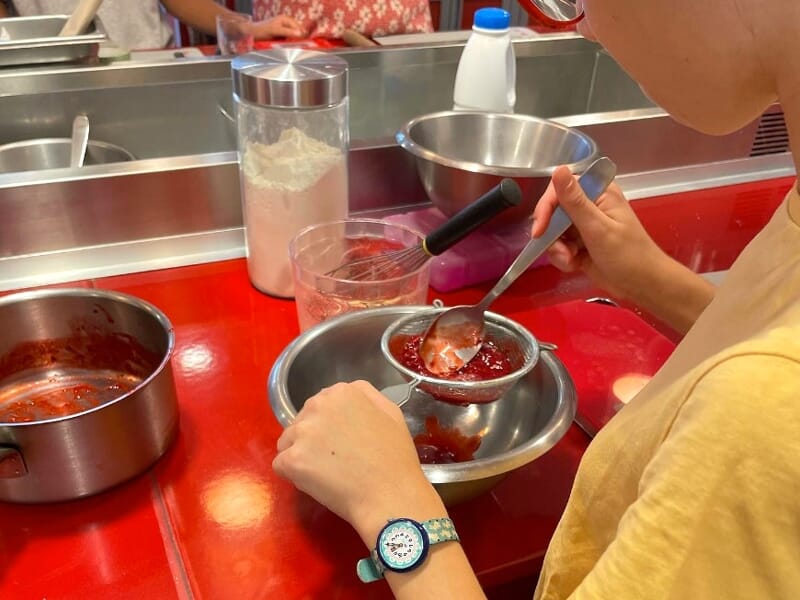 Une personne tamise une préparation rouge dans un bol en inox sur un plan de travail rouge dans une cuisine.