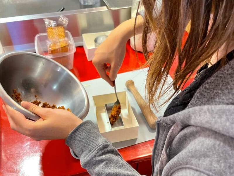 Une personne verse une préparation dans un moule carré blanc, tenant un bol en inox dans l'autre main.