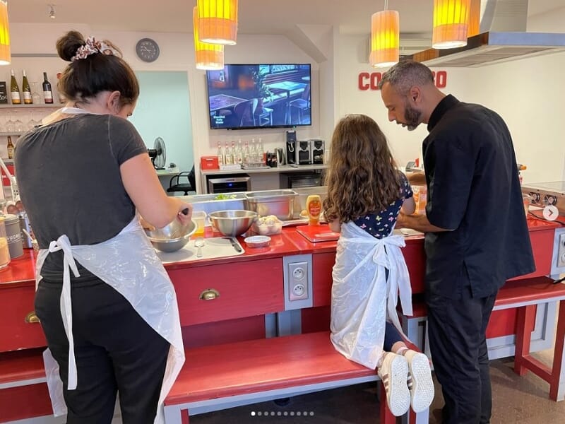 Trois personnes en tablier participent à un cours de cuisine dans une cuisine équipée avec un plan de travail rouge.