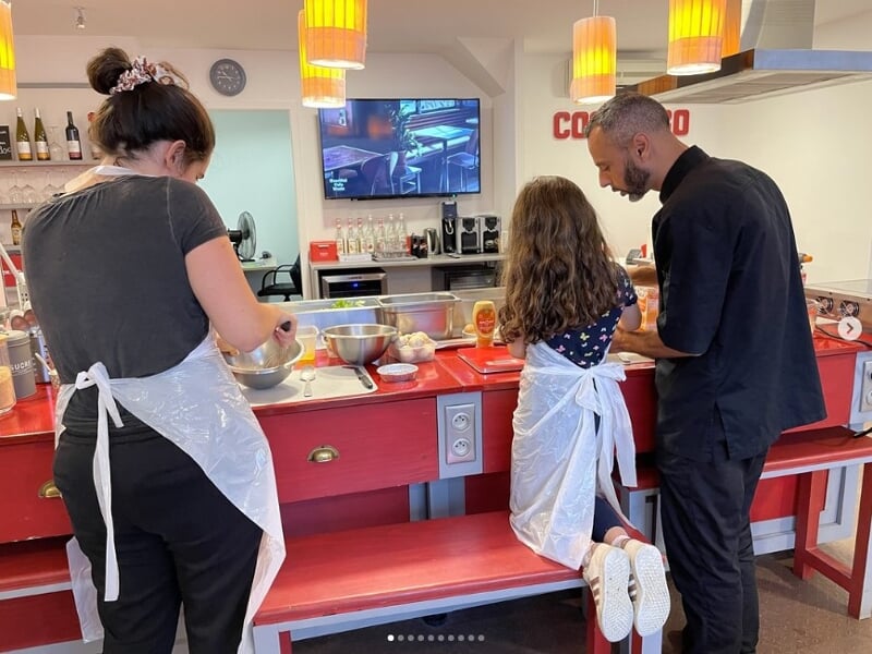 Trois personnes en tablier participent à un cours de cuisine dans une cuisine équipée avec un plan de travail rouge.