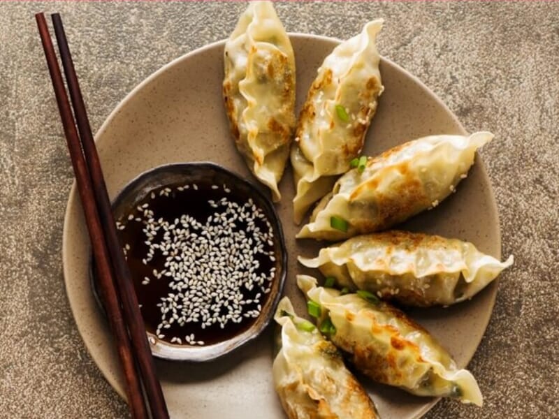 Assiette de gyozas dorés accompagnée d'une sauce soja aux graines de sésame, avec des baguettes en bois.
