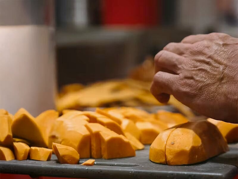 Main d'une personne coupant des morceaux de courge orange sur une planche dans une cuisine ou un atelier culinaire.