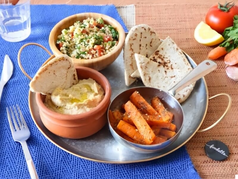 Assiette avec houmous, carottes rôties, pain pita et salade de couscous aux légumes frais sur table bleue.
