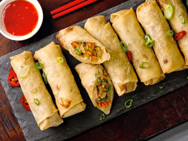 Rouleaux de printemps croustillants garnis de légumes, servis avec une sauce rouge épicée dans un bol blanc.
