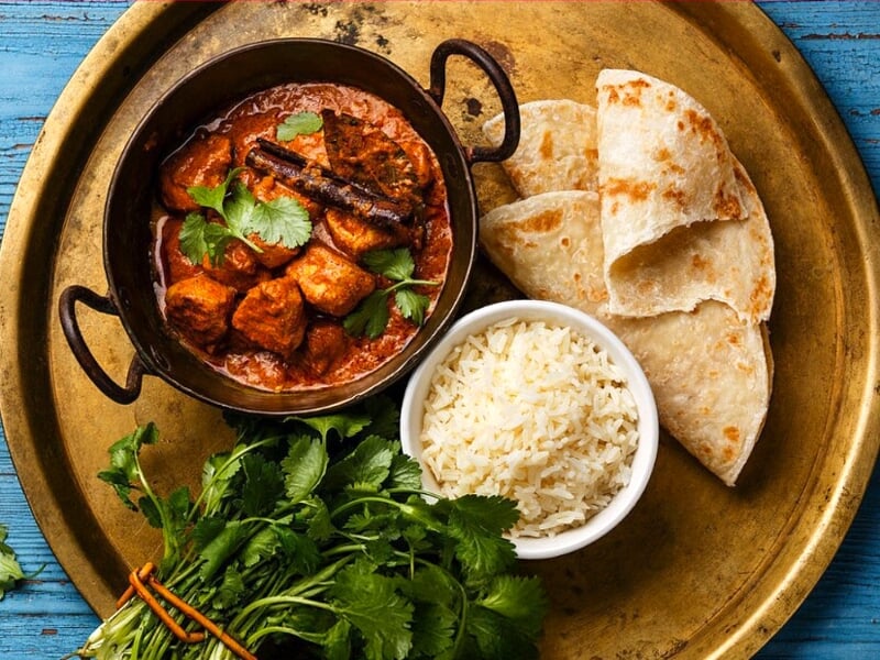 Plat indien avec curry de poulet, riz blanc, pain naan et coriandre fraîche sur plateau doré.
