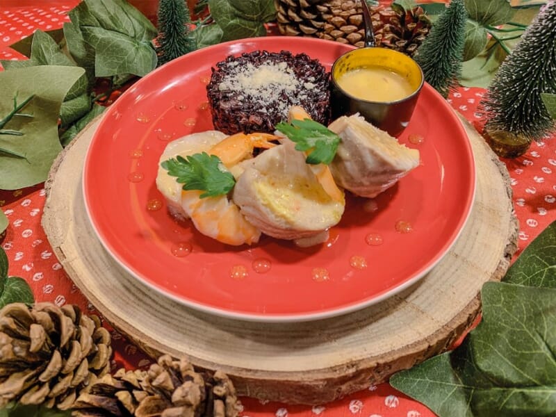 Assiette rouge avec crevettes, sauce, et accompagnement, décorations de Noël et pommes de pin autour.