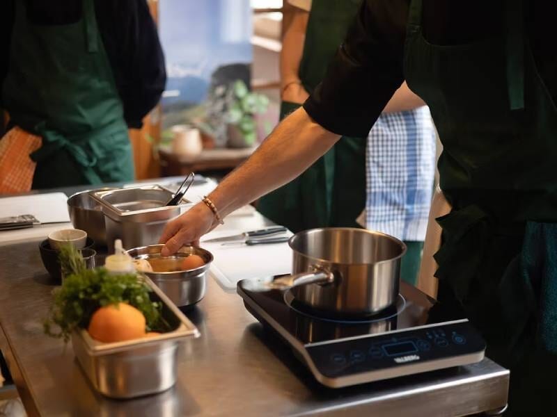 Personnes en tablier préparant un plat sur une plaque de cuisson électrique dans une cuisine bien équipée.